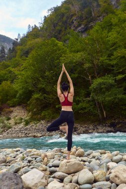 Doğada yoga dersleri. Tek başına spor yapma kavramı. Sosyal dışlama. Bir kadın kayalıklarda yoga yapıyor, bir dağ nehrinin yanında.