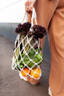 Kız elinde sebzeli örgü alışveriş poşeti, poşetsiz yeşillikler tutuyor. Sıfır atık, plastiksiz çevre dostu kavram. El yapımı makrome çanta. Sürdürülebilir yaşam tarzı.  