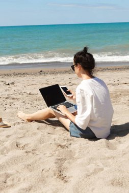 Dizüstü bilgisayar kullanan ve kumsalda cep telefonu tutan genç bir kadın. Tatil yaşam tarzı iletişimi. Serbest çalışma kavramı