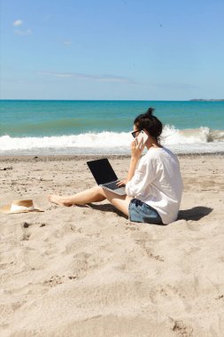 Genç bir kadın dizüstü bilgisayar kullanıyor ve sahilde telefonla konuşuyor. Tatil yaşam tarzı iletişimi. Serbest çalışma kavramı