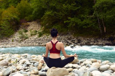 Doğada yoga dersleri. Tek başına spor yapma kavramı. Sosyal dışlama. Bir kadın kayalıklarda yoga yapıyor, bir dağ nehrinin yanında.