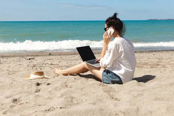 Genç bir kadın dizüstü bilgisayar kullanıyor ve sahilde telefonla konuşuyor. Tatil yaşam tarzı iletişimi. Serbest çalışma kavramı
