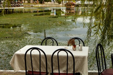 Gölün yanındaki kafede bir masa. Masayı beyaz bir masa örtüsü örtüyor ve dört sandalye yan yana duruyor.