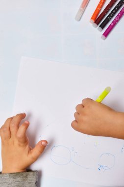 Sarı saçlı sevimli küçük çocuk evde renkli kalemler çiziyor. Beyaz masaya çiziyor. Beyaz kağıda resim çizen çocuğun ellerini kapat 