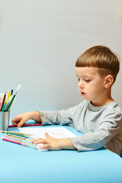 Милый маленький мальчик со светлыми волосами рисует цветные карандаши дома. Рисунки за синим столом.