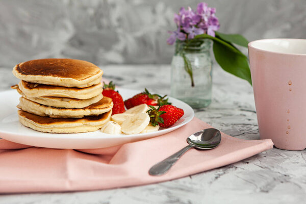 A stack of pancakes with a pink tea mug and  fresh strawberries on a white plate on a light background. A small bouquet of lilacs in a glass vase stands next to the Breakfast. 