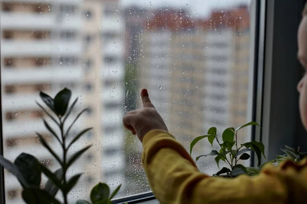 A little boy looks sadly out of the window. The concept of rainy weather. The child stays at home because of the rain