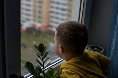 A little boy looks sadly out of the window. The concept of rainy weather. The child stays at home because of the rain clipart