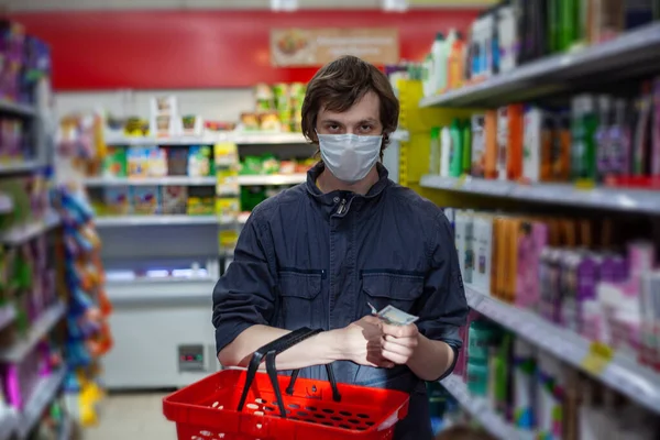 Man wears disposable face mask, makes shopping, thinks about lack of money during coronavirus pneumonia outbreak, poses with basket in department store. Concept of a crisis during an epidemic