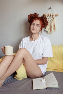 Very pretty redhead girl with dreads on bed in bedroom in the morning. A feminine, gentle girl. girl having breakfast in bed holds cup of coffee and reads book clipart
