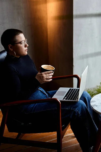 A young girl with glasses, short hair and a nose piercing is sitting in a cafe and working on a laptop. The concept of freelancing and remote work or training.