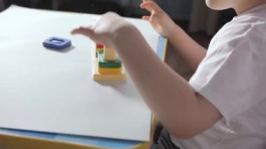 Beyaz Masa 'da oturan ve evde tahta ayırıcı ile oynayan bir çocuk. Beyaz tişörtlü bir çocuk eğitici oyunlar oynuyor. Mantığın gelişimi, şekiller ve renkler üzerine çalışma. Yüksek kaliteli FullHD görüntüler
