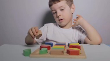 Beyaz Masa 'da oturan ve evde tahta ayırıcı ile oynayan bir çocuk. Beyaz tişörtlü bir çocuk eğitici oyunlar oynuyor. Mantığın gelişimi, şekiller ve renkler üzerine çalışma. Yüksek kaliteli FullHD görüntüler