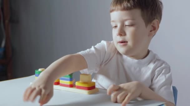 Chlapec sedí u bílého stolu a hraje si s dřevěným třídičem doma. Dítě v bílém tričku hraje vzdělávací hry. Vývoj logiky, studium tvarů a barev. Vysoce kvalitní FullHD záběry
