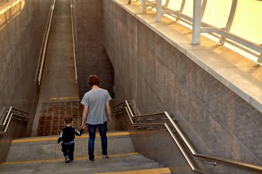 Modern yeraltı geçidi. Bir adam ve bir çocuk merdivenlerden aşağı iniyor. Arkadan bak. Ön planda adımlar ve tırabzanlar. 
