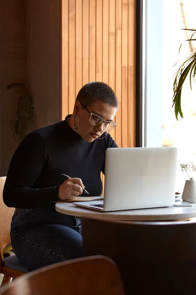 A young girl with glasses, short hair and a nose piercing is sitting in a cafe and working on a laptop. The concept of freelancing and remote work or training.
