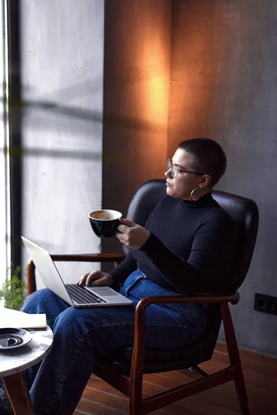 A young girl with glasses, with short hair and a nose piercing is sitting in a cafe, drinks coffee and working on a laptop. The concept of freelancing and remote work or training.