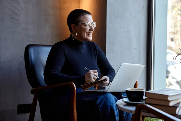 A young girl with glasses, with short hair is sitting in a cafe and working on a laptop. The girl looks at her laptop and makes notes in a notebook. The concept of distance learning