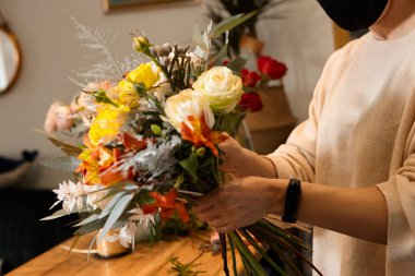 Çiçekçi renklerle çalışır. Çiçekçi gelecekteki buket için çiçek seçiyor. Çiçekçi dükkanında maske takmış vazodaki çiçekleri kontrol ediyor..