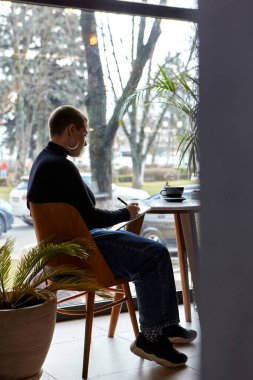 Kısa saçlı ve burun piercingli genç bir kız bir kafede oturuyor ve tablette çalışıyor. Serbest çalışma ve uzak çalışma kavramı..