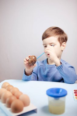 Küçük bir çocuk masaya mavi bir arka plan üzerine paskalya yumurtası çiziyor. Küçük çocuk evde boyalı Paskalya yumurtalarını boyuyor. Çocukların Paskalya yaratıcılığı. Paskalya yumurtalarını bir çocuk süslüyor..
