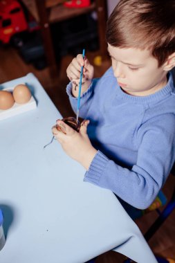 Küçük bir çocuk masaya mavi bir arka plan üzerine paskalya yumurtası çiziyor. Küçük çocuk evde boyalı Paskalya yumurtalarını boyuyor. Çocukların Paskalya yaratıcılığı. Paskalya yumurtalarını bir çocuk süslüyor..