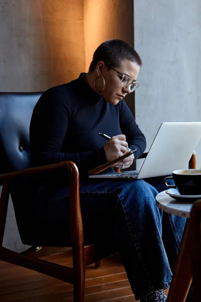 A young girl with glasses, with short hair is sitting in a cafe and working on a laptop. The girl looks at her laptop and makes notes in a notebook. The concept of distance learning