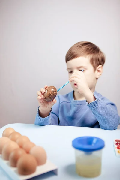 一个小孩在蓝色的背景上 在桌子旁边画了一个复活节彩蛋 小男孩在家里画复活节彩蛋 儿童的复活节创造力 孩子装饰复活节彩蛋 — 图库照片