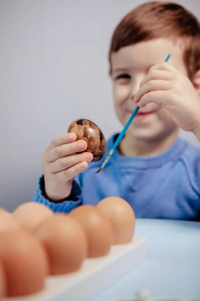 一个小孩在蓝色的背景上 在桌子旁边画了一个复活节彩蛋 小男孩在家里画复活节彩蛋 儿童的复活节创造力 孩子装饰复活节彩蛋 — 图库照片