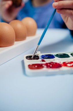 Küçük bir çocuk masaya mavi bir arka plan üzerine paskalya yumurtası çiziyor. Küçük çocuk evde boyalı Paskalya yumurtalarını boyuyor. Çocukların Paskalya yaratıcılığı. Paskalya yumurtalarını bir çocuk süslüyor..