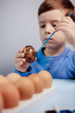 Küçük bir çocuk masaya mavi bir arka plan üzerine paskalya yumurtası çiziyor. Küçük çocuk evde boyalı Paskalya yumurtalarını boyuyor. Çocukların Paskalya yaratıcılığı. Paskalya yumurtalarını bir çocuk süslüyor..