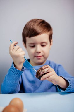 Küçük bir çocuk masaya mavi bir arka plan üzerine paskalya yumurtası çiziyor. Küçük çocuk evde boyalı Paskalya yumurtalarını boyuyor. Çocukların Paskalya yaratıcılığı. Paskalya yumurtalarını bir çocuk süslüyor..