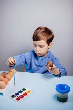 Küçük bir çocuk masaya mavi bir arka plan üzerine paskalya yumurtası çiziyor. Küçük çocuk evde boyalı Paskalya yumurtalarını boyuyor. Çocukların Paskalya yaratıcılığı. Paskalya yumurtalarını bir çocuk süslüyor..