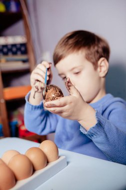 Küçük bir çocuk masaya mavi bir arka plan üzerine paskalya yumurtası çiziyor. Küçük çocuk evde boyalı Paskalya yumurtalarını boyuyor. Çocukların Paskalya yaratıcılığı. Paskalya yumurtalarını bir çocuk süslüyor..