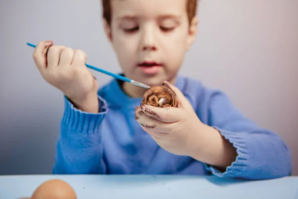 一个小孩在蓝色的背景上 在桌子旁边画了一个复活节彩蛋 小男孩在家里画复活节彩蛋 儿童的复活节创造力 孩子装饰复活节彩蛋 — 图库照片