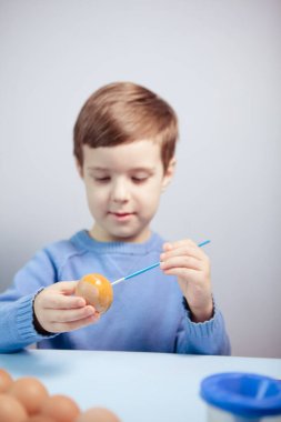 Küçük bir çocuk masaya mavi bir arka plan üzerine paskalya yumurtası çiziyor. Küçük çocuk evde boyalı Paskalya yumurtalarını boyuyor. Çocukların Paskalya yaratıcılığı. Paskalya yumurtalarını bir çocuk süslüyor..