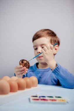 Küçük bir çocuk masaya mavi bir arka plan üzerine paskalya yumurtası çiziyor. Küçük çocuk evde boyalı Paskalya yumurtalarını boyuyor. Çocukların Paskalya yaratıcılığı. Paskalya yumurtalarını bir çocuk süslüyor..