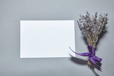 Beyaz kağıt kaplamalı lavanta çiçekleri gri arka planda. Beyaz kağıt boş, gri masada kurumuş çiçekler. Davetiye kartı mockup.Düz konum, üst görünüm, kopyalama alanı, maket