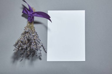 Beyaz kağıt kaplamalı lavanta çiçekleri gri arka planda. Beyaz kağıt boş, gri masada kurumuş çiçekler. Davetiye kartı mockup.Düz konum, üst görünüm, kopyalama alanı, maket