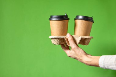Siyah kapaklı iki bardak kahverengi kağıt tutan eller. İki özel kahve ya da tanıtım. Yeşil arka planda iki bardak tutan eller. Çay ya da kahve. Siyah kapaklı kahverengi kağıt bardak..