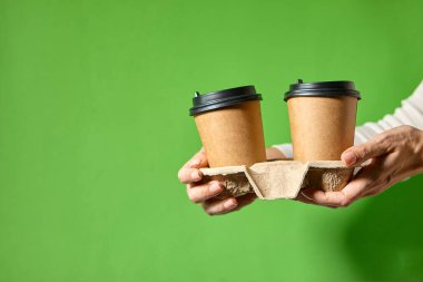 Siyah kapaklı iki bardak kahverengi kağıt tutan eller. İki özel kahve ya da tanıtım. Yeşil arka planda iki bardak tutan eller. Çay ya da kahve. Siyah kapaklı kahverengi kağıt bardak..
