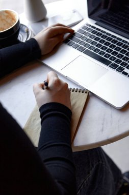 Genç bir kız bir kafede oturuyor, dizüstü bilgisayarla çalışıyor ve not defterine not alıyor. Yakın çekim, ellerin fotoğrafı. Serbest çalışma ve uzak çalışma veya eğitim kavramı.
