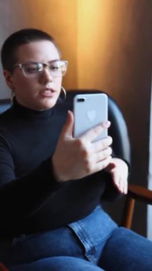 Kısa saçlı ve burun piercingli genç bir kız bir kafede oturup telefonundaki video bağlantısıyla konuşuyor. Kız canlı yayın ya da yayın yapıyor.