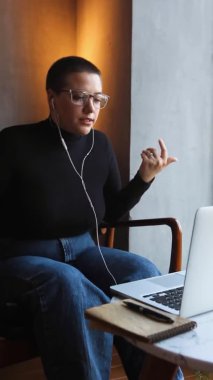 Kulaklık takan, kısa saçlı ve burun piercingli genç bir kız bir kafede oturuyor ve laptopun yanındaki video bağlantısıyla konuşuyor.