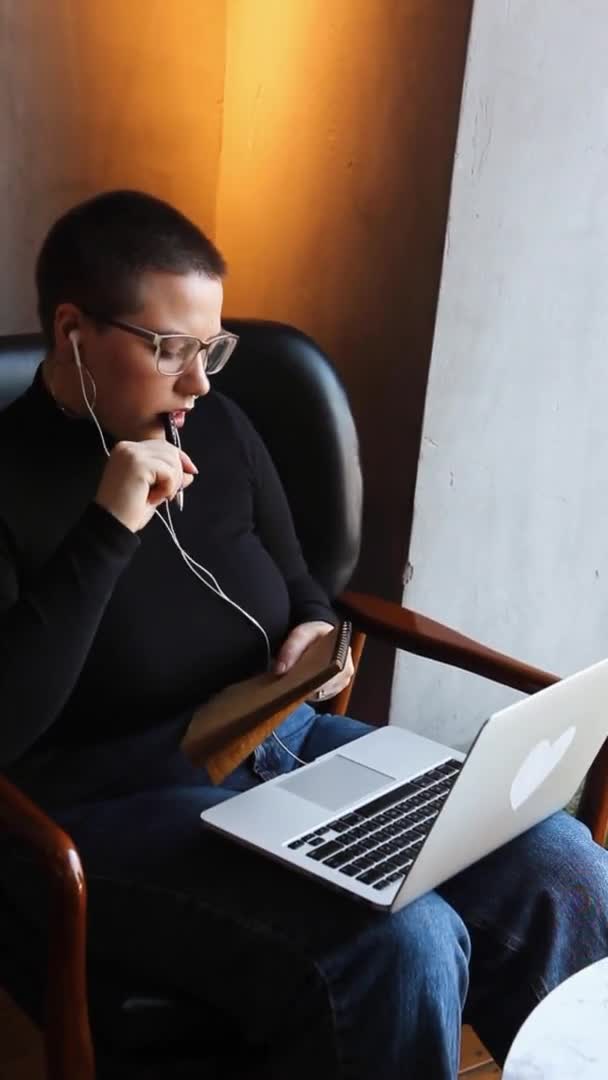 一个戴眼镜 留着短发的年轻女孩坐在咖啡店里 在笔记本电脑上工作 这个女孩看了看她的笔记本电脑 在笔记本上做了笔记 远距离学习的概念 — 图库视频影像