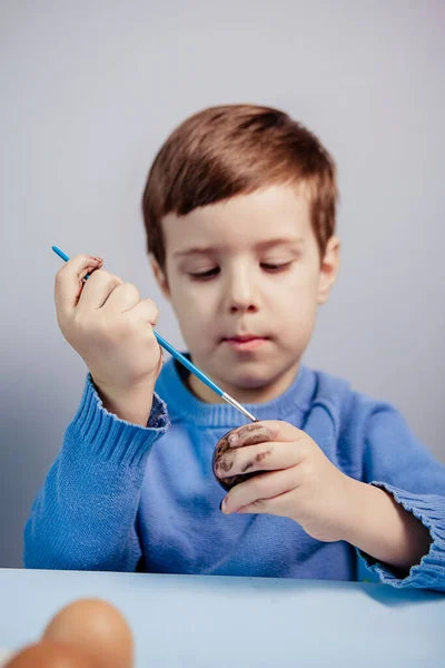 一个小孩在蓝色的背景上 在桌子旁边画了一个复活节彩蛋 小男孩在家里画复活节彩蛋 儿童的复活节创造力 孩子装饰复活节彩蛋 — 图库照片