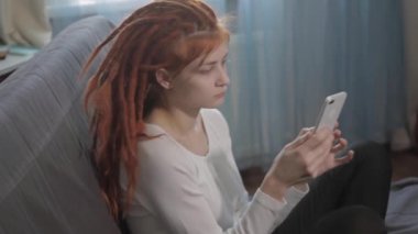 Rastalı kızıl saçlı bir kız evinde kanepede oturmuş kitap okuyor ve akıllı telefonundan gelen çağrıya cevap veriyor.