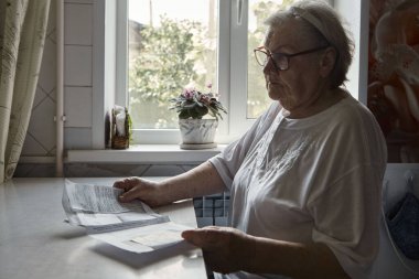Evdeki mutfaktaki masada oturan yaşlı kadın evdeki faturaları tutuyor, ödemeyi veya borç oluşturmayı unuttuğundan endişeleniyor. 80 'li yaşlarda emekli bir kadın.
