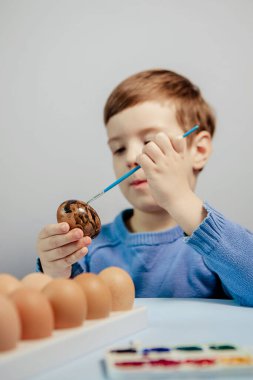 Küçük bir çocuk masaya mavi bir arka plan üzerine paskalya yumurtası çiziyor. Küçük çocuk evde boyalı Paskalya yumurtalarını boyuyor. Çocukların Paskalya yaratıcılığı. Paskalya yumurtalarını bir çocuk süslüyor..
