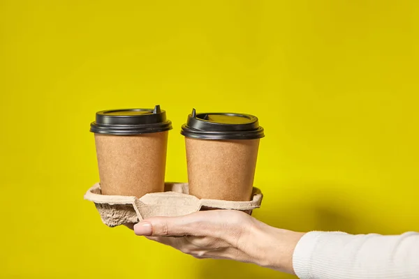 Hands holding two cups brown paper with black lid. Two coffee special offer or promo. Hands holding two cups on yellow screen background. Tea or Coffee to go. Brown paper cup with black lid.